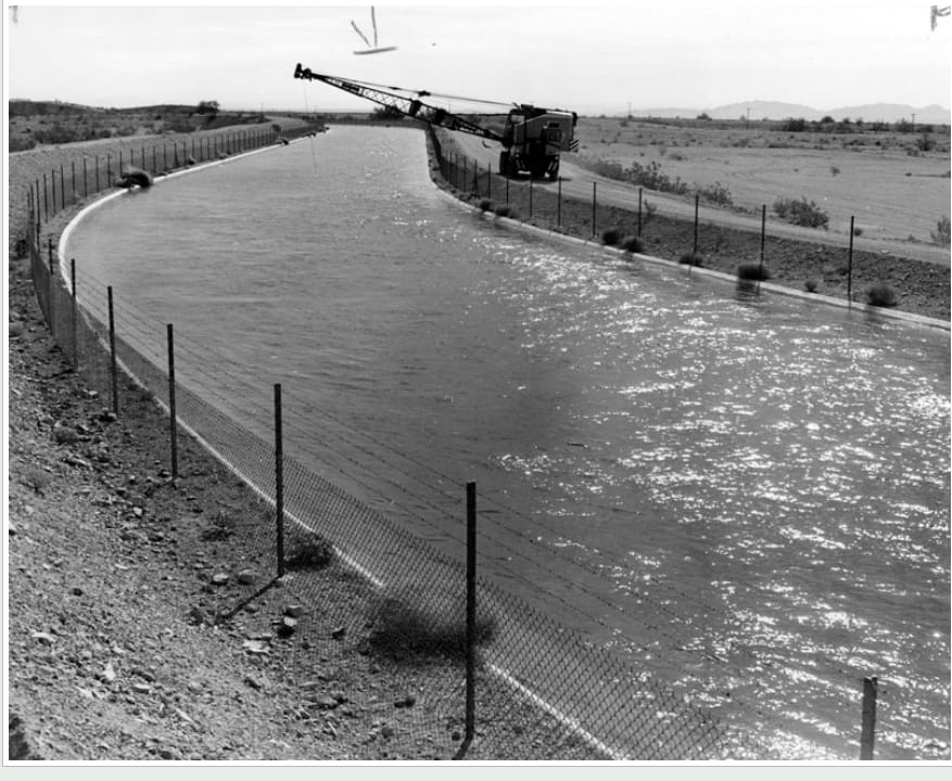 A 1977 photograph of the concrete aqueduct canal filled to the brim with sparkling water, a dragline crane parked on the far bank.