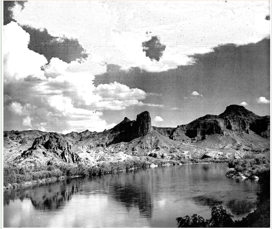 A 1930s photograph of the Colorado River winding between rugged desert mountains under a cloud-filled sky.