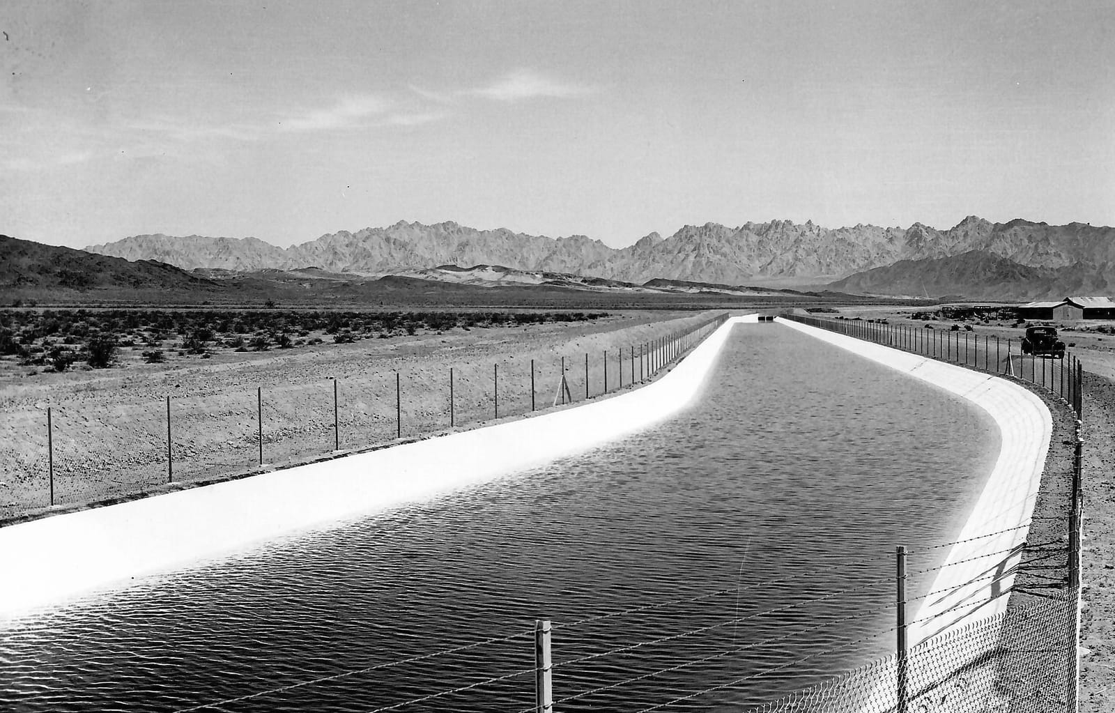 A wide concrete-lined canal filled with water curving through flat desert toward a jagged mountain range, fenced on both banks.