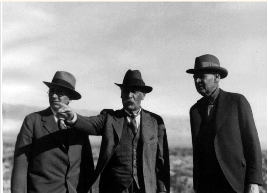 Three men in suits and hats standing on open desert ground during a 1931 survey trip, one gesturing toward the horizon.