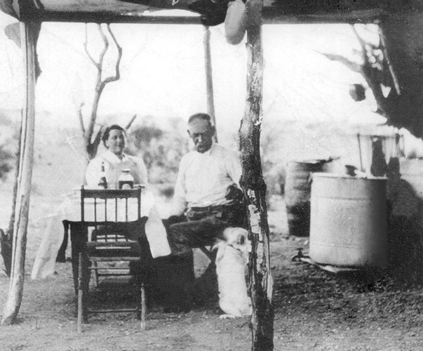 A black-and-white photograph from 1906 showing Wyatt Earp and his wife Josephine seated in the shade of a brush ramada at their desert mining camp, with a water barrel and camp furnishings around them.