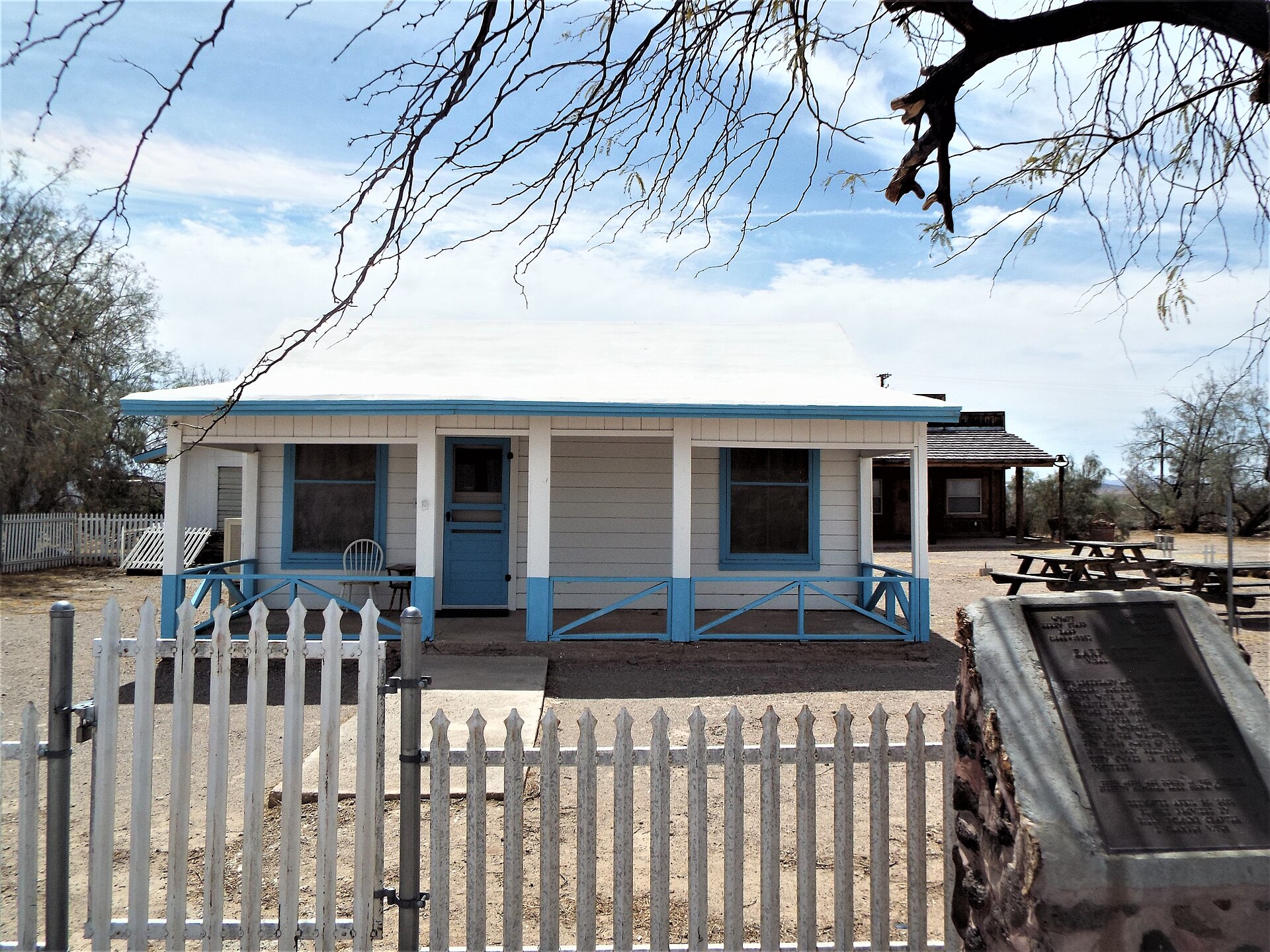 The Earp Cottage today: a small white clapboard house with sky-blue trim and a shaded front porch, set behind a white picket fence, with a bronze memorial plaque on a stone base at right.