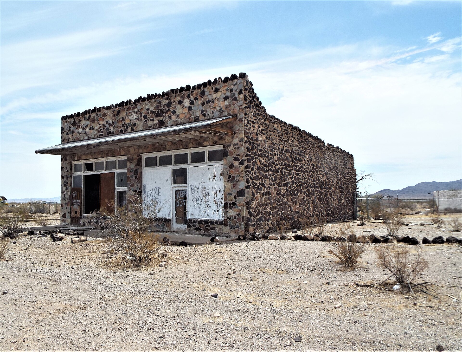 The J. M. Heacock Building today: a single-story commercial building faced entirely in rounded desert fieldstone and mortar, with a shaded storefront porch, boarded windows, and open desert and mountains behind it.
