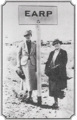Two women in 1930s dress standing beside a desert signpost reading 'EARP.'