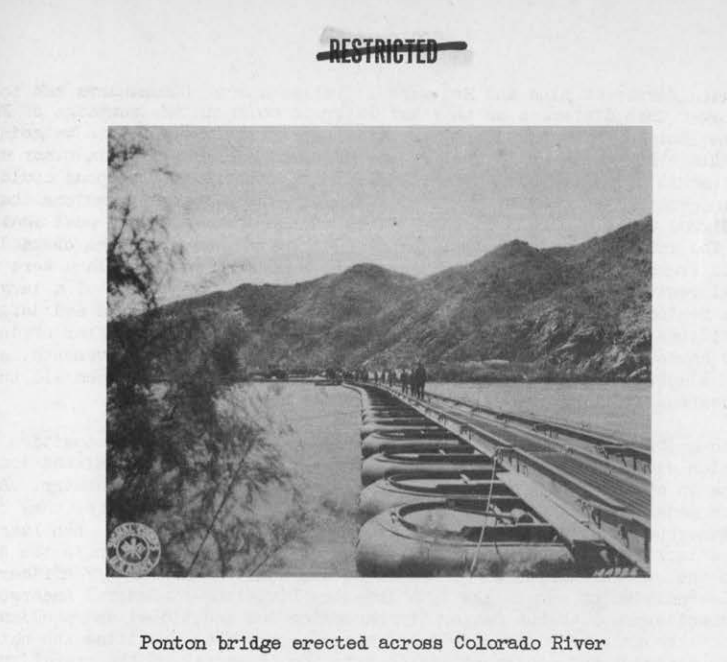 A pontoon bridge on inflated floats stretching across the Colorado River toward rugged mountains, with soldiers crossing on foot.