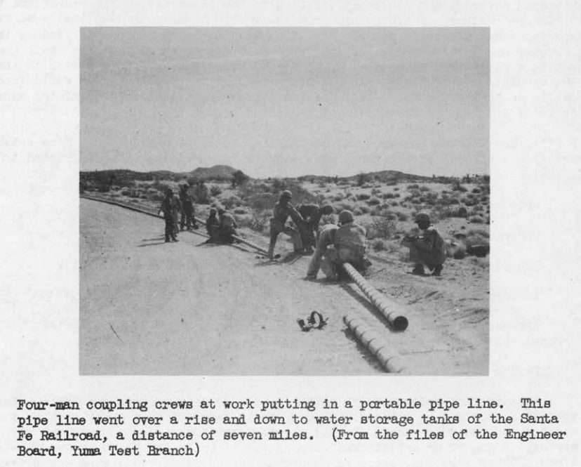 Soldiers in helmets working in crews to couple sections of a portable pipeline laid along a desert road.