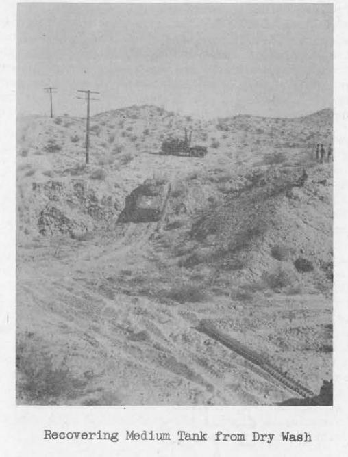 A medium tank being winched up a steep, rocky desert wash by a recovery vehicle at the crest of the slope.