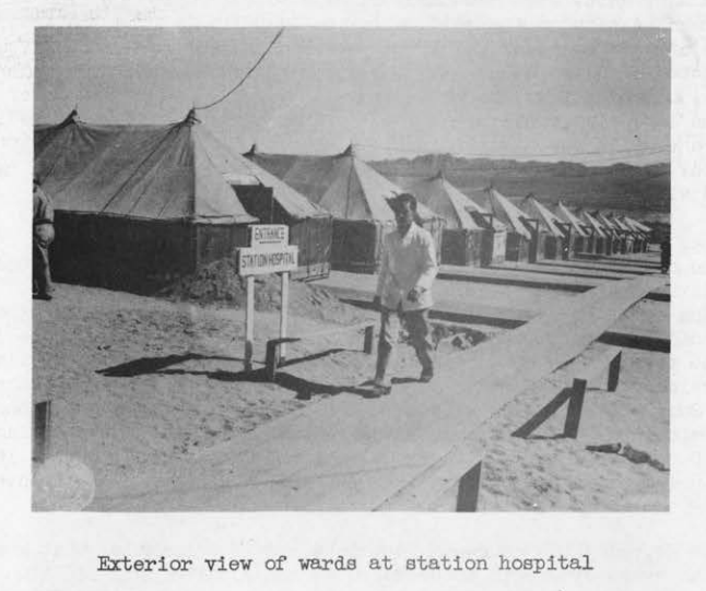 A long row of pyramidal hospital tents pitched on the desert, with a soldier walking past a sign reading Entrance Station Hospital.