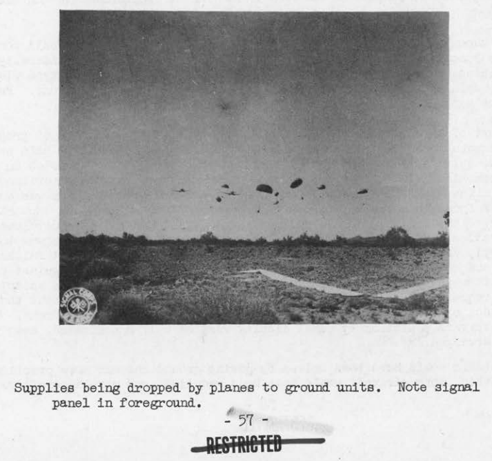 Several parachutes carrying supply bundles descending toward open desert scrub, with a signal panel laid out on the ground below.