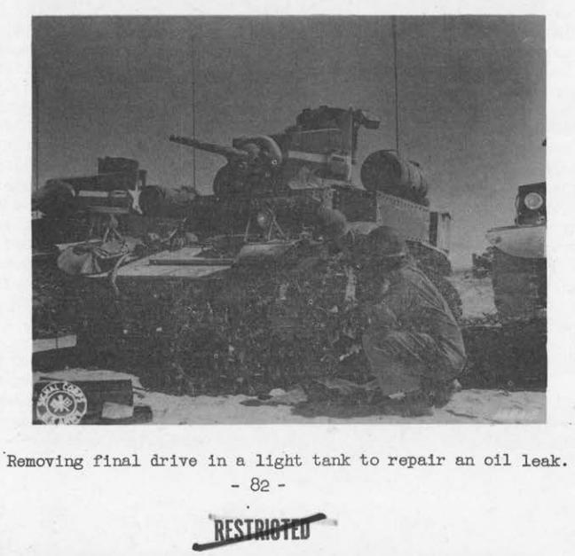 A soldier crouched at the front of a light tank in the desert, working to remove its final drive assembly.