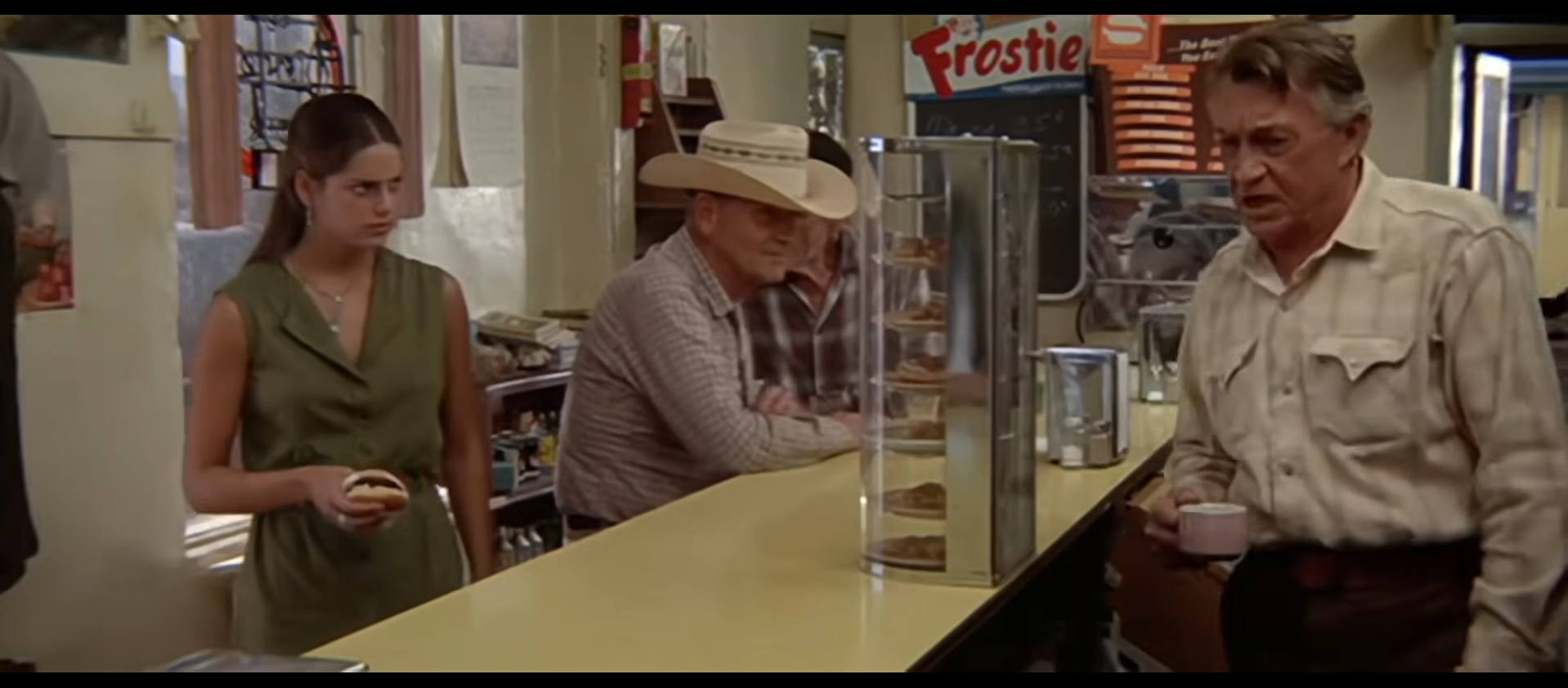 A film still from Zabriskie Point inside a desert cafe: a young woman at a yellow counter holding a roll, an older man in a white cowboy hat, and a gray-haired man with a coffee cup, beneath 'Frostie' and 'Squirt' signs.