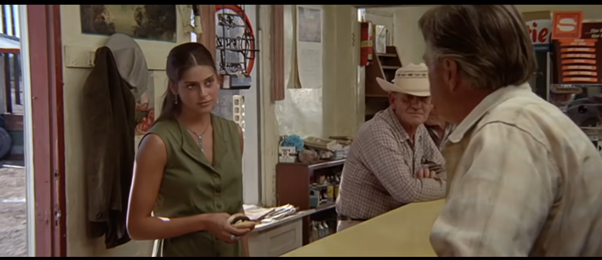 A film still from Zabriskie Point: the young woman in an olive dress speaks across the cafe counter, with the man in the white cowboy hat seated behind her.