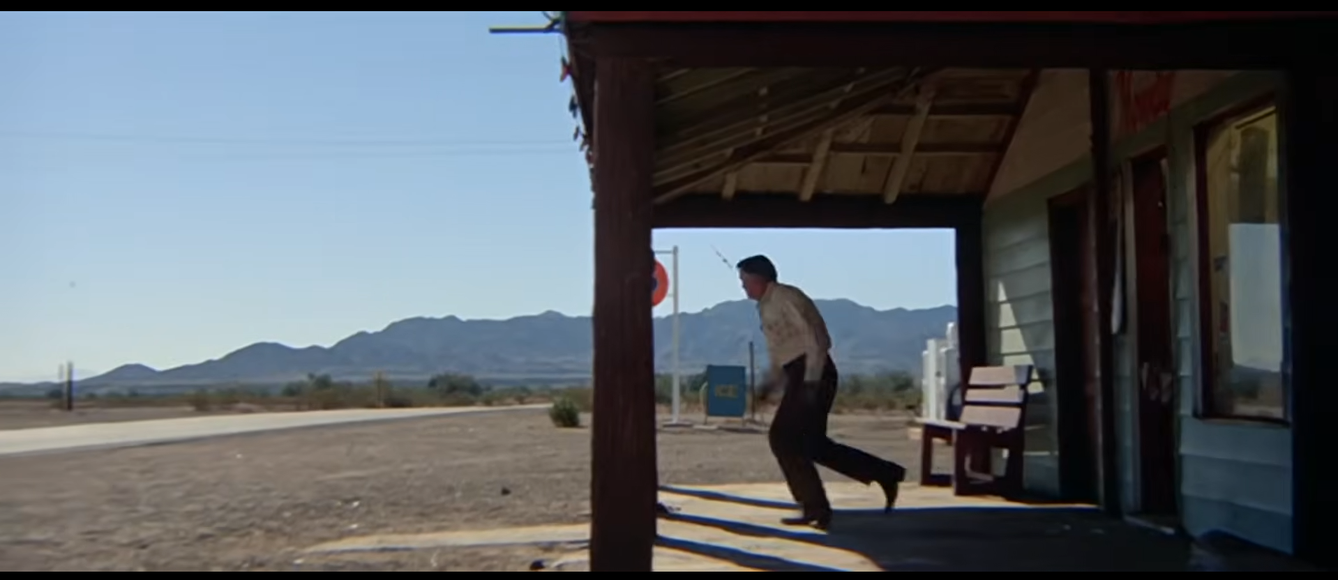A film still from Zabriskie Point: a man bounds off the shaded wooden porch of a desert store into bright daylight, mountains on the horizon behind him.