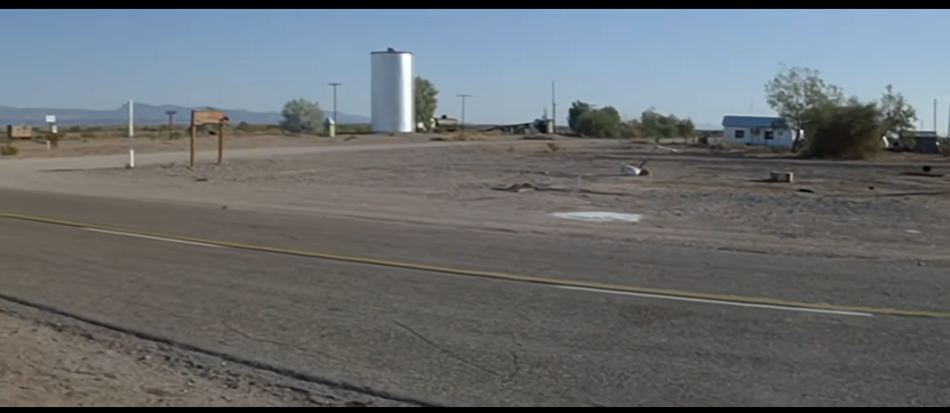 A film still from Zabriskie Point: a wide view of the Vidal townsite along the highway, with a white water tank, a small blue building, and open desert beyond.
